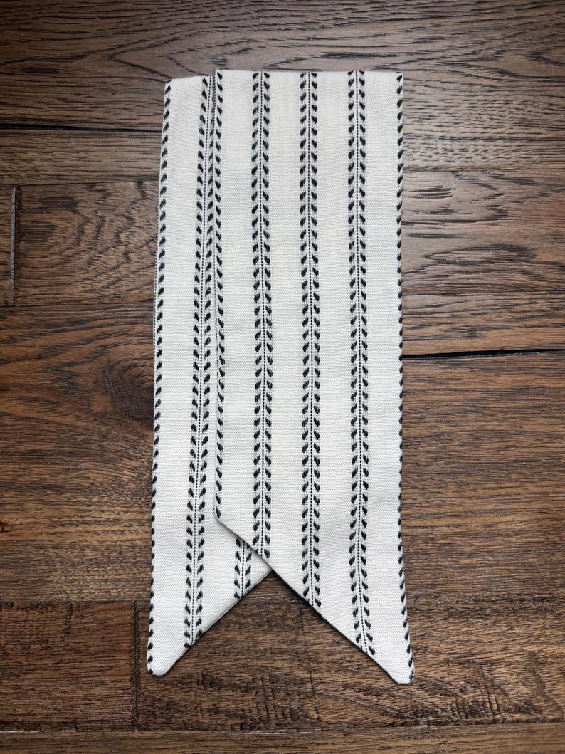 May include: A long, rectangular fabric scarf in an off-white color featuring vertical stripes of black, repeating leaf-like patterns. The scarf has pointed, triangular ends that overlap slightly, creating a clean and symmetrical appearance.