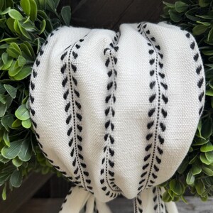 May include: A white fabric ribbon featuring a repeating black vertical leaf or vine pattern. The ribbon is gathered and draped in soft folds, showcasing a textured weave. It is positioned against a background of dense, dark green foliage.