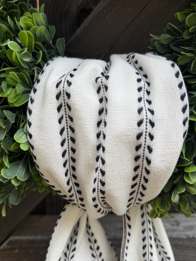 May include: A white fabric ribbon featuring a repeating black vertical leaf or vine pattern. The ribbon is gathered and draped in soft folds, showcasing a textured weave. It is positioned against a background of dense, dark green foliage.