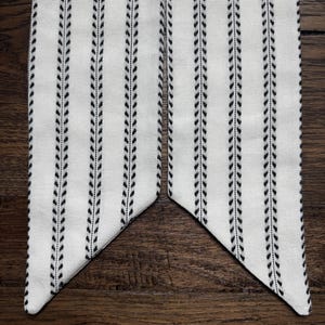 May include: A long, narrow fabric strip featuring a white background with vertical black stripes composed of small, repeating leaf-like patterns. The ends of the fabric are cut at an angle, creating a pointed shape on each side of the strip.