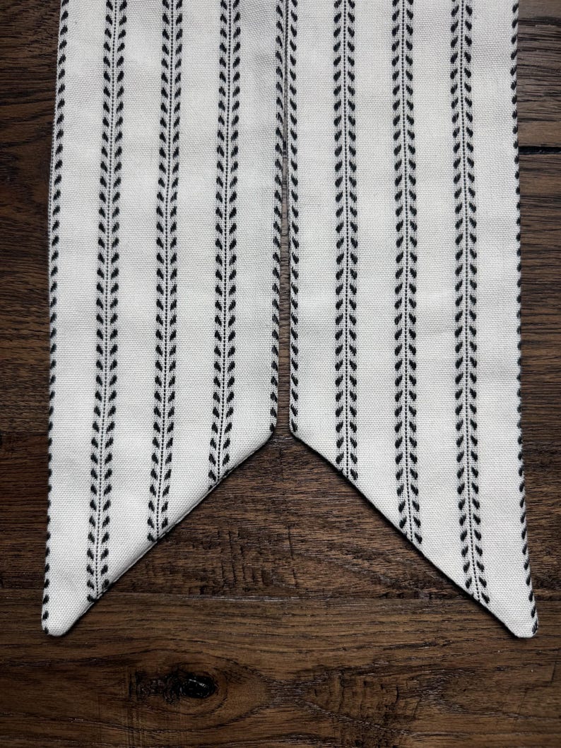 May include: A long, narrow fabric strip featuring a white background with vertical black stripes composed of small, repeating leaf-like patterns. The ends of the fabric are cut at an angle, creating a pointed shape on each side of the strip.