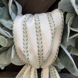 May include: A decorative wreath featuring soft, sage green lamb's ear leaves arranged in a circular pattern. A cream-colored fabric ribbon with vertical olive green leaf-patterned stripes is tied in a knot at the center, with two long tails hanging down.
