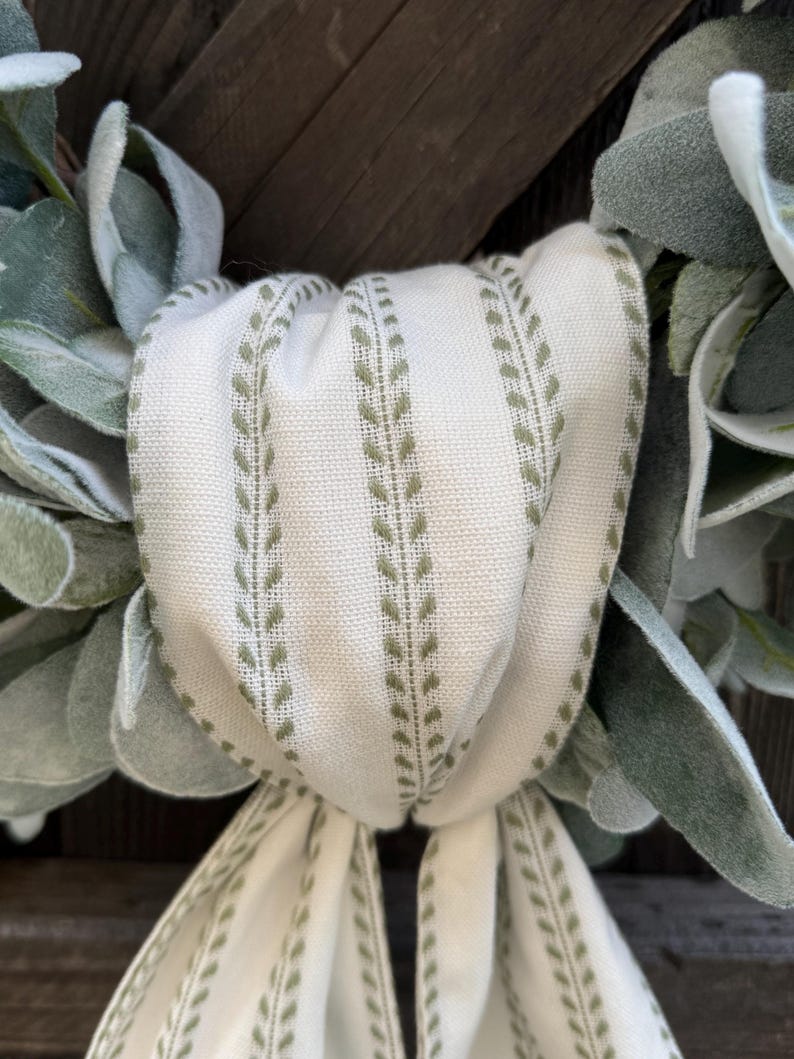 May include: A decorative wreath featuring soft, sage green lamb's ear leaves arranged in a circular pattern. A cream-colored fabric ribbon with vertical olive green leaf-patterned stripes is tied in a knot at the center, with two long tails hanging down.