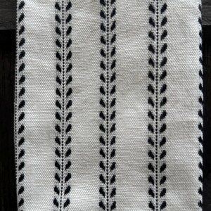 May include: A woven fabric featuring a repeating pattern of vertical black stripes on an off-white background. Each stripe consists of a central line flanked by small, symmetrical leaf-like shapes, creating a consistent botanical-inspired design.