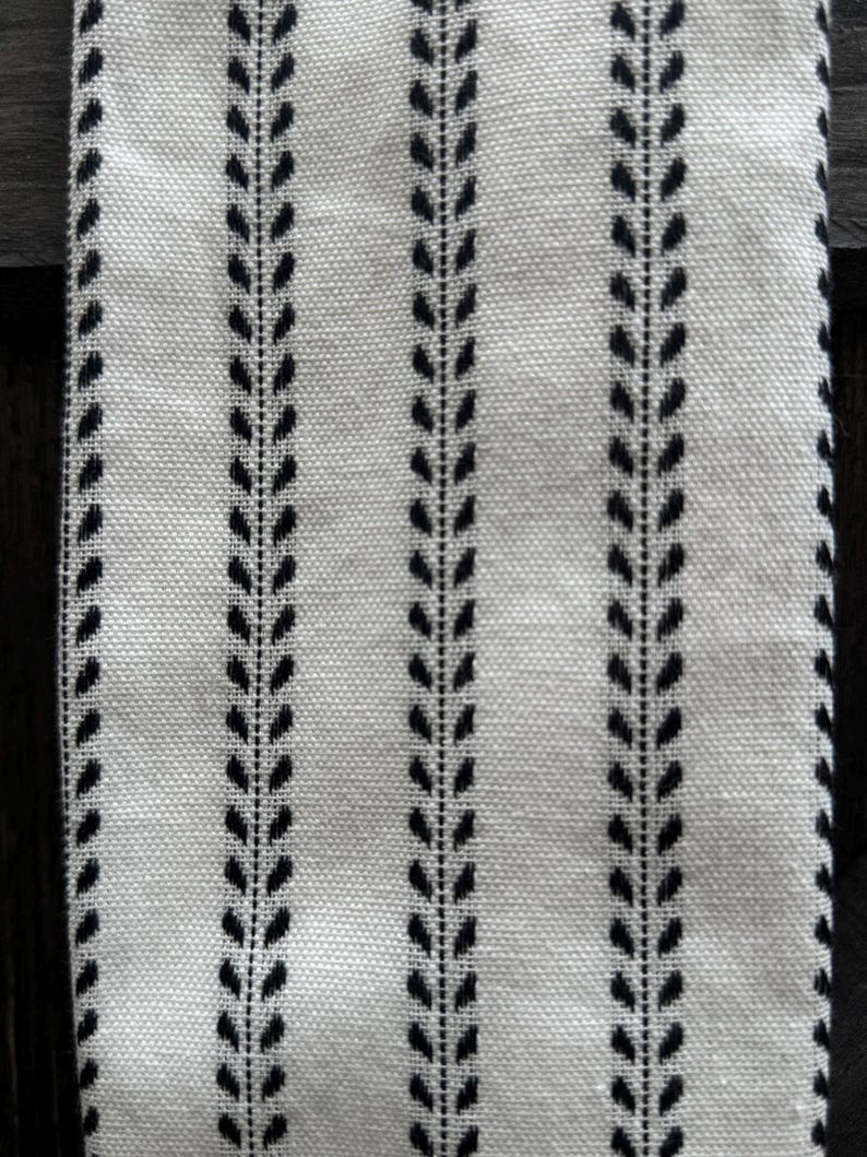 May include: A woven fabric featuring a repeating pattern of vertical black stripes on an off-white background. Each stripe consists of a central line flanked by small, symmetrical leaf-like shapes, creating a consistent botanical-inspired design.