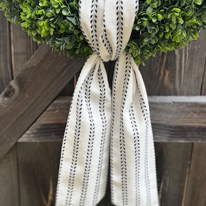 May include: A green boxwood wreath adorned with a long, off-white fabric ribbon featuring a repeating black vertical leaf-like pattern. The ribbon is tied in a simple knot at the top center of the circular wreath, with two long ends hanging down.