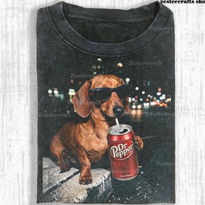 May include: Dark gray t-shirt featuring a brown dachshund wearing sunglasses and drinking from a Dr. Pepper can. The dog is set against a blurred city lights background. The text "zesteecrafts shop" is visible.