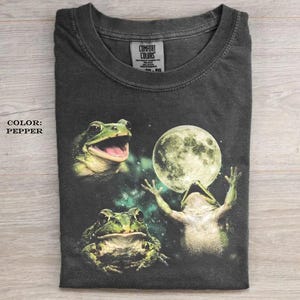 May include: A charcoal gray t-shirt featuring a graphic print of three frogs and a full moon against a dark, nebulous background. The shirt has a crew neckline and a Comfort Colors brand label on the inside collar. The text COLOR: PEPPER is visible.