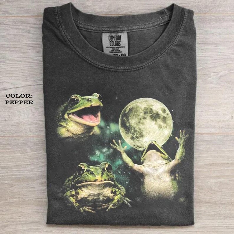 May include: A charcoal gray t-shirt featuring a graphic print of three frogs and a full moon against a dark, nebulous background. The shirt has a crew neckline and a Comfort Colors brand label on the inside collar. The text COLOR: PEPPER is visible.