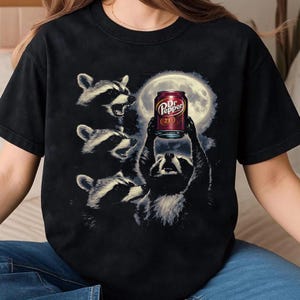 May include: Black t-shirt featuring a graphic of raccoons gathered around a can of Dr. Pepper. The can is held up in the air against a full moon backdrop. The raccoons are in various poses, with detailed fur.