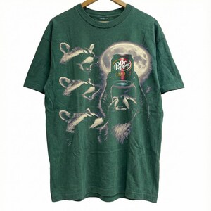May include: Green t-shirt featuring a graphic of raccoons and a Dr. Pepper can in front of a full moon. The raccoons are in various poses, with one holding the soda can. The Dr. Pepper can has a red and white label.
