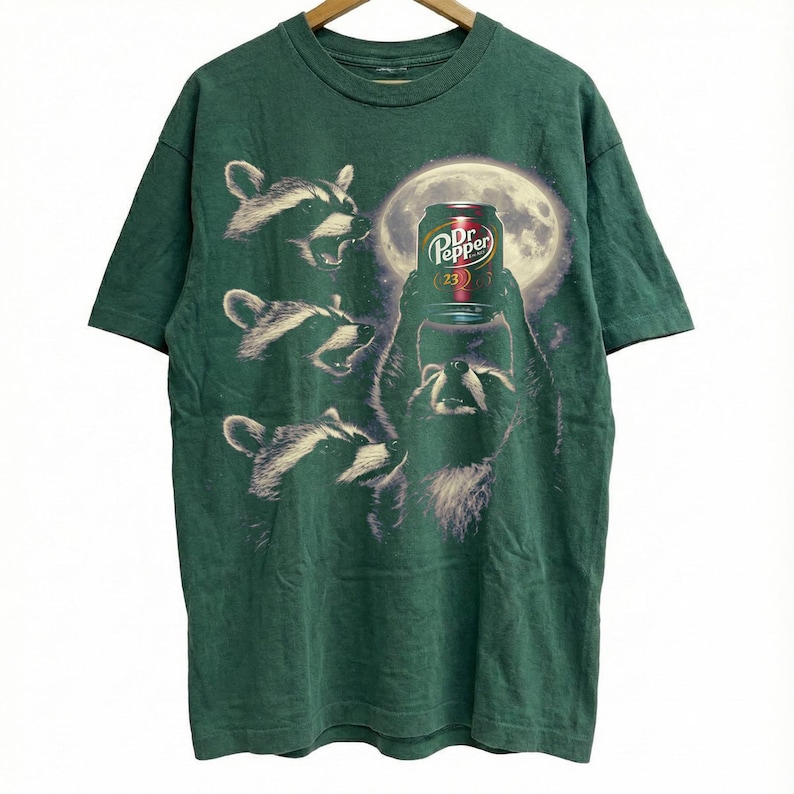 May include: Green t-shirt featuring a graphic of raccoons and a Dr. Pepper can in front of a full moon. The raccoons are in various poses, with one holding the soda can. The Dr. Pepper can has a red and white label.