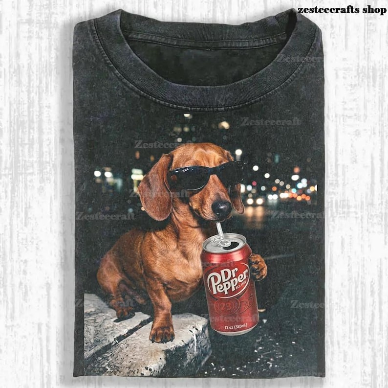 May include: Dark gray t-shirt featuring a brown dachshund wearing sunglasses and drinking from a Dr. Pepper can. The dog is set against a blurred city lights background. The text "zesteecrafts shop" is visible.