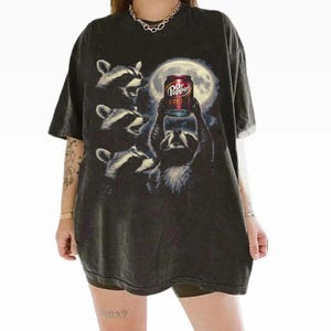 May include: Dark gray t-shirt featuring a graphic of raccoons gathered around a full moon, with one raccoon holding a Dr. Pepper can. The Dr. Pepper can has the number 23 on it. The shirt has a vintage, distressed look.