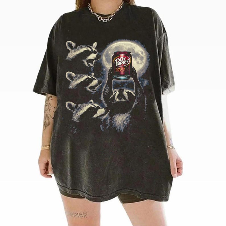 May include: Dark gray t-shirt featuring a graphic of raccoons gathered around a full moon, with one raccoon holding a Dr. Pepper can. The Dr. Pepper can has the number 23 on it. The shirt has a vintage, distressed look.