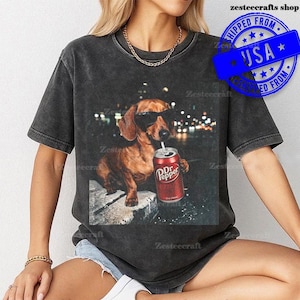 May include: Dark gray t-shirt featuring a brown dachshund wearing sunglasses and drinking from a Dr. Pepper can. The dog is sitting on a stone surface with a blurred city background. The shirt has the text "ZesteeCrafts Shop" and a "SHIPPED FROM USA" stamp.