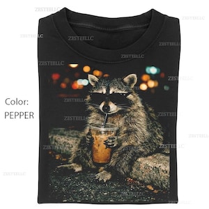 May include: Black t-shirt featuring a raccoon wearing sunglasses and holding an iced coffee. The raccoon is sitting on a stone surface with a blurred background of city lights. The shirt is pepper-colored.