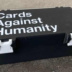 以下が含まれることがあります： 「Cards Against Humanity」の白文字が入った黒い長方形の箱。前面には白いプラスチック製の留め金が付いています。カードゲームを収納するように設計された箱です。