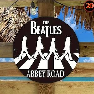 Può includere: Un cartello rotondo in metallo nero con la scritta bianca "THE BEATLES" e una silhouette bianca della band su un attraversamento pedonale. Sotto, "ABBEY ROAD". Il cartello è montato su una struttura in legno, con una lampadina appesa.