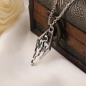 May include: A silver-toned metal pendant featuring an intricate, stylized dragon emblem enclosed within a diamond-shaped frame. The pendant hangs from a matching silver-toned link chain, showcasing a polished finish with dark recessed detailing.