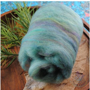 May include: A thick, cylindrical roll of unspun wool roving featuring a variegated blend of soft teal, mint green, and subtle lavender tones. The fibre has a fluffy, airy texture with visible wisps and a soft, matte appearance throughout the roll.