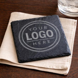 May include: Square black slate coaster with the text "YOUR LOGO HERE" in a circular design. The coaster rests on a beige linen napkin, with a dark wood background. A glass of water is visible in the upper right corner.