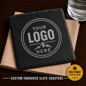 May include: A square, black slate coaster with a custom logo design. The logo is in a circular frame with the text "YOUR LOGO HERE" and a mountain graphic. The coaster rests on a tan napkin. Text at the bottom reads "CUSTOM ENGRAVED SLATE COASTERS".