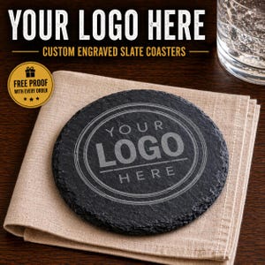 May include: A round black slate coaster with a rough, natural edge featuring an engraved circular design with the text YOUR LOGO HERE in the center. The coaster is placed on a folded beige fabric napkin, with additional text above.