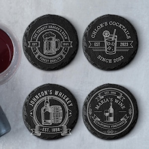 May include: Four round black slate coasters with white engraved designs. One coaster features a beer mug, another a cocktail, a third a whiskey bottle and barrel, and the last a wine bottle and glass. Each coaster has text.
