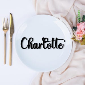 May include: A white ceramic dinner plate featuring the name Charlotte in black cursive script lettering, positioned next to a gold-toned metal fork and knife, with a cluster of pink artificial roses resting on a soft, draped fabric surface.