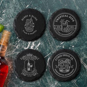 May include: Four black slate coasters with white engraved designs. One coaster reads "Berry & Bliss" with a jar of berries. Another says "Tropical Sips" with a cocktail. The third says "Chateau Noble" with a wine bottle and glass. The last says "Citrus Grove Co."