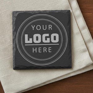 May include: Square black slate coaster with the text "YOUR LOGO HERE" in a white circular design. The coaster is resting on a beige linen napkin, with a wooden surface in the background.