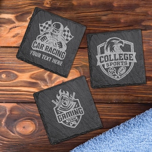 May include: Three square black slate coasters with engraved designs. One features a race car driver with the text "CAR RACING YOUR TEXT HERE". Another has a college sports emblem with an eagle, basketball, and football. The third coaster says "GAMING TEAM".