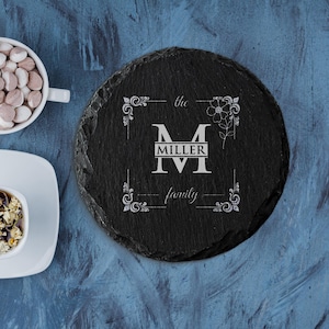 May include: A black slate coaster with a decorative border and the text "the MILLER family". The coaster has a rough, natural edge and a dark, textured surface. A white floral design is also present.