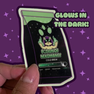 Può includere: Un adesivo con un design di sacchetto di caffè nero con la scritta "Potion of Nekromancy Cold Brew". L'adesivo ha un effetto verde che si illumina al buio e un'illustrazione di un gatto dei cartoni animati. È presente anche la scritta "Glows in the Dark!".