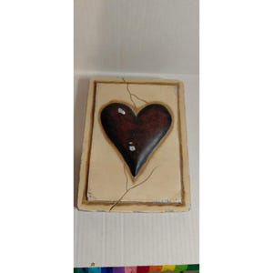 May include: A rectangular, off-white decorative plaque with a dark red heart in the center. The heart has a glossy finish and is slightly raised. A crack runs through the plaque, adding a rustic touch.