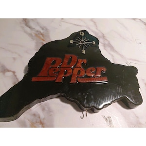 May include: A vintage Dr. Pepper sign with a clock and two hooks. The sign is black with the Dr. Pepper logo in red and brown. The clock has a gold-colored face and hands. The sign is shaped like a state.