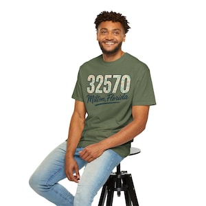 May include: Olive green t-shirt with the text "32570 Milton, Florida" in a vintage style. The shirt is worn by a person sitting on a black stool. The text is in a white and green floral pattern.