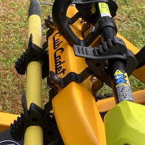 May include: A yellow Cub Cadet equipment rack mounted on a machine, featuring black rubberized clamps holding a yellow pole and a black pole with warning labels. The assembly includes metal brackets and hardware for securing outdoor tools.