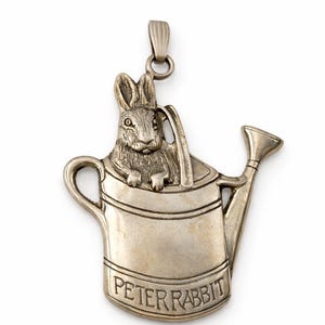 May include: A silver-toned pendant featuring a rabbit inside a watering can. The rabbit is detailed with visible ears and eyes. The watering can has a handle and spout. The words "PETER RABBIT" are inscribed on the can.