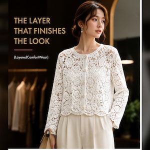 Op de afbeelding: Een wit vest met lange mouwen van kant, een ronde halslijn en een knoopsluiting aan de voorzijde. De tekst op de afbeelding luidt: THE LAYER THAT FINISHES THE LOOK (LayeredComfortWear). Het kledingstuk heeft een bloemrijk haakpatroon.