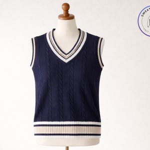 Puede incluir: Chaleco de punto trenzado azul marino con cuello en V y rayas blancas, beige y azul marino. El chaleco se muestra en un maniquí. La imagen incluye un logotipo circular que dice "Sweater & Vest" y "Knit to Impress".
