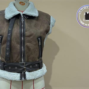 Peut inclure: Un gilet en daim marron avec un col et une bordure en fausse fourrure blanche. Le gilet est doté d'une fermeture éclair sur le devant, de deux poches zippées et d'une ceinture à la taille. Un logo "Sweater & Vest" est en arrière-plan.