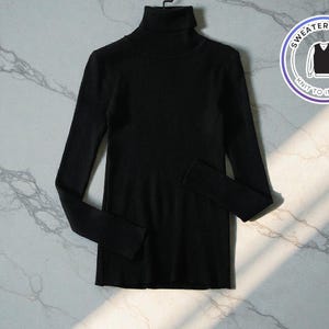 Peut inclure: Un pull noir à col roulé avec des manches longues, présenté sur un cintre. Le pull a une texture côtelée et une coupe ajustée. Un logo circulaire avec le texte "SWEATER & VEST" et un graphique de gilet est visible.