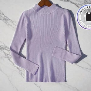 May include: A light purple ribbed knit sweater with long sleeves and a mock turtleneck. The sweater is displayed on a wooden hanger against a marble background. A circular graphic with the text "SWEATER & VEST" and "KNIT TO IMPRESS" is in the upper right corner.