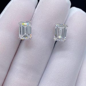 May include: Two emerald-cut diamond stud earrings. The diamonds are set in a white metal, likely platinum or white gold. The earrings are displayed against a white background, creating a contrast that highlights the brilliance of the diamonds.