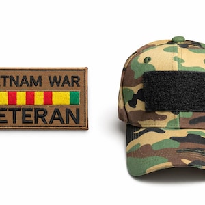 Könnte beinhalten: Eine Camouflage-Baseballkappe mit einem schwarzen Aufnäher und einem braunen Aufnäher mit dem Text "VIETNAM WAR VETERAN" und einem farbenfrohen Streifendesign. Die Kappe ist in Grün-, Braun- und Beigetönen gehalten.
