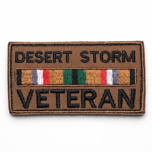 Może przedstawiać: Prostokątna brązowa naszywka z czarnym napisem "DESERT STORM VETERAN". Posiada centralny pasek z kolorowymi paskami: szarym, białym, czerwonym i zielonym, reprezentującymi wstążkę służby wojskowej.