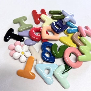 May include: A selection of brightly coloured ceramic letters and a flower. The letters are in various colours, including red, blue, green, and yellow. The flower is white with a pink centre. The letters are in a variety of fonts and sizes.