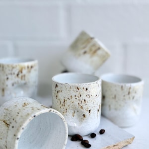 May include: A collection of four ceramic cups featuring a white base with brown speckled patterns and subtle indentations on the sides. The cups are arranged in a cluster, with one cup lying on its side and several dark roasted coffee beans scattered.